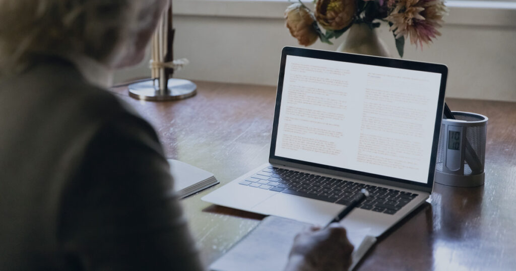 Person viewing text on a laptop while taking handwritten notes at a desk.